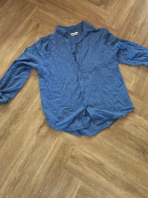 Blå skjorta i linne  - Säljer en enkel blå skjorta med knappar framtill och klassisk krage. Skjortan har lång ärm och är tillverkad i linne perfekt för en avslappnad stil. Passformen är normal och tyget är enfärgat utan mönster.