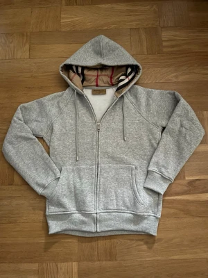 Grå zip hoodie med mönstrad huva - Säljer en grå hoodie med dragkedja och fickor framtill. Huvan har ett snyggt mönstrat innerfoder i beige, svart och rött. Tröjan har ribbade muddar vid ärmslut och nederkant samt dragsko i huvan. Perfekt för en chill och avslappnad stil.