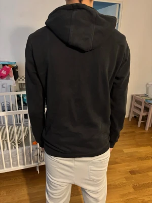 Svart hoodie från Adidas - Säljer en svart hoodie från Adidas med klassisk vit logga på bröstet. Hoodien har huva, känguruficka framtill och ribbade muddar vid ärmar och nederkant. Perfekt för en chill och sportig look.