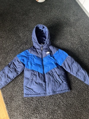 Blå pufferjacka från Nike 158-170/ M - Säljer en blå pufferjacka från Nike i storlek 158-170. Jackan har två nyanser av blått med en snygg Nike swoosh detalj över bröstet, dragkedja framtill och huva. Perfekt för kalla dagar och riktigt skön att ha på sig. Materialet är syntetiskt och jackan har två sidofickor. Köpt på JD sports!