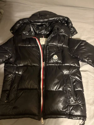 Svart dunjacka från Moncler - Snygg svart dunjacka från Moncler med glansig finish, huva och dragkedja med rödvit detalj. Jackan har två fickor med dragkedja, en bröstficka med Moncler-logga och klassisk serietidningsetikett på insidan. Perfekt för kalla dagar.