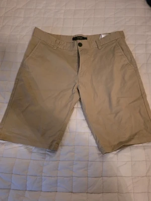Beige shorts från SMOG - Snygga beige shorts från SMOG med klassisk passform och två sidofickor. Perfekta för varma dagar och enkla att matcha med olika stilar. Tillverkade i mjuk bomull för skön komfort.