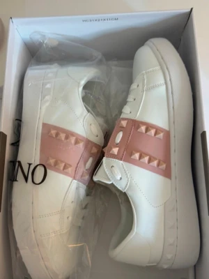 Vita sneakers med rosa nitar - Snygga vita sneakers med bred rosa rem över mitten, dekorerad med pyramidformade nitar i matchande rosa. Skorna har klassisk låg profil, vita snören och detaljerad sula. Tillverkade i slätt läderliknande material för en clean look.