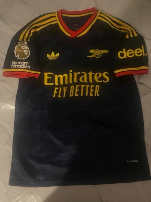 Arsenal matchtröja svart/gul - Snygg Arsenal matchtröja i svart med gula detaljer och röda inslag. Tröjan har klubbmärke, Adidas-logga och sponsortryck på bröstet samt Premier League-märke på ärmen. Tillverkad i lätt och ventilerande material, perfekt för fotboll eller träning.