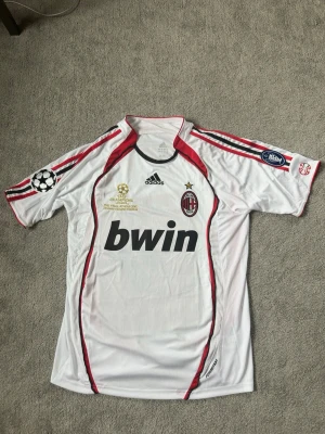 AC Milan Champions League tröja - Vit AC Milan matchtröja från Adidas med röda och svarta detaljer. Champions League 2007-finaltryck i guld på bröstet, klubbmärke och Adidas-logga. Svart 'bwin' sponsortryck på magen och officiella turneringsmärken på ärmarna. Klassisk sportig passform i funktionsmaterial. !NYSKICK!