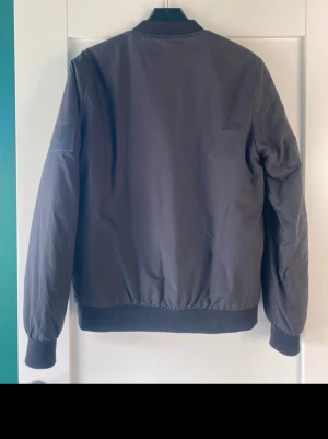 Svart bomberjacka Peak Performance - Snygg svart bomberjacka från Peak Performance i klassisk modell. Jackan har ribbade muddar vid ärmslut och midja samt diskret logga på ärmen. Tillverkad i ett slitstarkt material som ger en clean och stilren look.