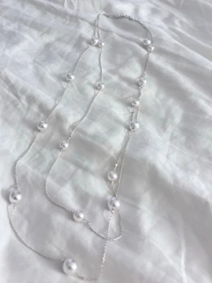 Silverhalsband med vita pärlor - Säljer ett elegant halsband med tunn silverfärgad kedja och vita pärlor jämnt utspridda längs hela halsbandet. Halsbandet har justerbar längd med förlängningskedja och karbinlås. Aldrig använt!