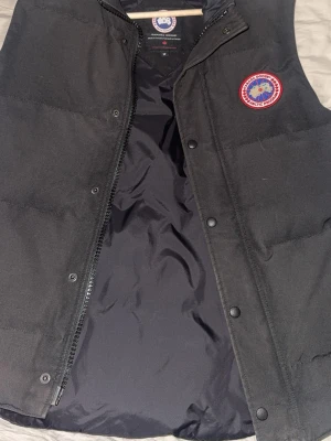 Svart dunväst från Canada Goose - Snygg svart dunväst från Canada Goose med klassisk logga på bröstet. Västen har dragkedja och tryckknappar framtill samt mjukt foder. Perfekt för lager-på-lager och håller dig varm under kyliga dagar. Säljs pga ligger bara i garderoben o dammar kommer inte till någon användning 