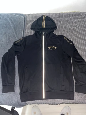 Svart hoodie från BOSS med dragkedja - Snygg svart hoodie från BOSS med gulddetaljer och dragkedja framtill. Huva med BOSS-logga på snöret och dekorativa sömmar på axlarna. Klassisk passform och två fickor framtill. Perfekt för en stilren och avslappnad look.