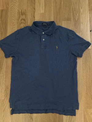 Ralph Lauren piketröja - Klassisk blå pikétröja från Polo Ralph Lauren med korta ärmar och krage. Tröjan har två knappar framtill och den ikoniska broderade loggan på bröstet. Perfekt till jeans eller shorts för en avslappnad look.