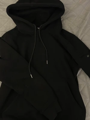 Svart hoodie från C.P. Company - Säljer en svart hoodie från C.P. Company med klassisk huva och dragsnören. Ärmen har en ikonisk linsdetalj och ficka med knapp. Hoodien har magficka och ribbade muddar. Perfekt för dig som gillar streetwear och stilren design.