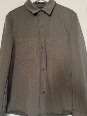 Grå overshirt från Zara - Stilren grå overshirt från Zara med två stora bröstfickor och knappar framtill. Skjortan har klassisk krage, lång ärm och är gjord i ett mjukt, strukturerat tyg som ger en avslappnad look.