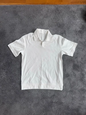Vit ribbad piké med kort ärm - Snygg vit piké med korta ärmar och diskret ribbad struktur. Klassisk krage och mjukt material som känns skönt mot huden. Perfekt för en clean look.