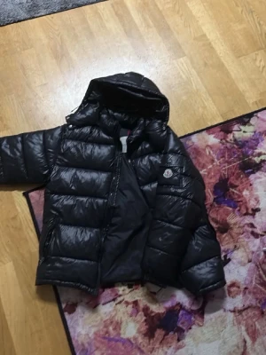 Svart dunjacka från Moncler - Säljer en svart dunjacka från Moncler med glansig finish och huva. Jackan har breda, quiltade paneler och dragkedja framtill samt två sidofickor. Klassisk Moncler-logga på ärmen. Perfekt för kalla vinterdagar. Har nfc tag
