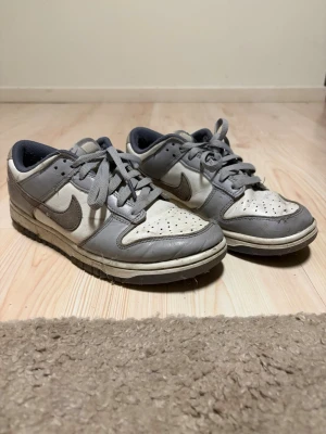 Gråvita Nike Dunk Low sneakers - Säljer ett par klassiska Nike Dunk Low sneakers i grått och vitt. Skorna har en låg siluett, perforerad tåbox och ikoniska swoosh-loggan på sidan. Ovandelen är i läder med matchande grå snören och vit mellansula. Perfekta för dig som gillar retrostil.