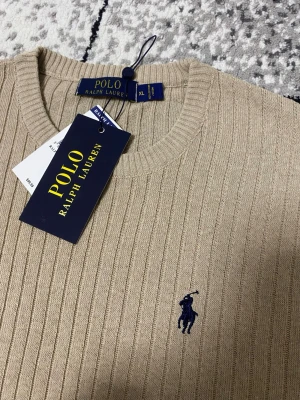 Beige stickad tröja Polo Ralph Lauren - Snygg beige stickad tröja från Polo Ralph Lauren med ribbad struktur och klassisk rund halsringning. Tröjan har det ikoniska blå broderade logotypen på bröstet och är perfekt för en stilren look. Den är ”halv armad” om man säger så för den är varken lång eller kort armad. Den är helt ny och jätte fin passar med flera saker. Står Xl men är i storlek S ungefär. Det är en tunn stickad tröja