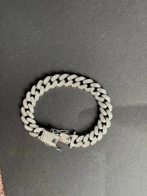 Iced ut cuban länkarmband silver - Snyggt cuban länkarmband i silverfärg med glittrande stenar över hela länkarna. Armbandet har en robust låsdetalj och breda länkar som ger en lyxig och trendig känsla. Perfekt för dig som vill sticka ut med accessoarer som syns.
