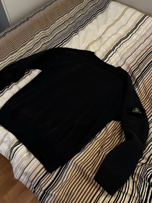 Svart sweatshirt från Stone Island - Säljer en svart sweatshirt från Stone Island med klassisk rund hals och ribbade muddar. Tröjan har den ikoniska Stone Island-loggan på vänster ärm och är tillverkad i mjukt material som passar perfekt till streetwear-stilen.