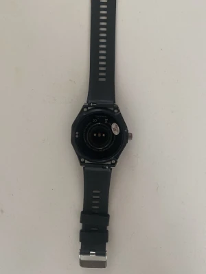Svart smartklocka med silikonarmband - Snygg svart smartklocka med rund urtavla och robust boett i metall. Klockan har ett svart silikonarmband med justerbara hål och metallspänne. Perfekt för dig som vill ha koll på träning och notiser direkt på handleden.