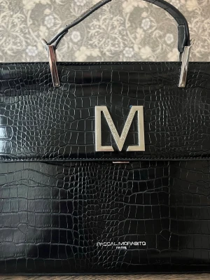 Pascal Morabito 'Magda Croco' Handväska – Svart Krokodilpräglad - Stilren svart handväska i croco-mönstrat konstläder från Pascal Morabito. Väskan har ett stort silverfärgat M på locket och eleganta metallhandtag. Perfekt accessoar för dig som gillar lyxig och trendig design. 33x26x12 cm