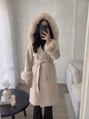 Pälskappa med knytskärp  - Säljer en elegant beige kappa med äkta päls på huva och ärmslut. Kappan har en knytning i midjan och är perfekt för kyligare dagar. Den har en mjuk och lyxig känsla, vilket gör den till ett stilrent val för höst och vinter. S/M