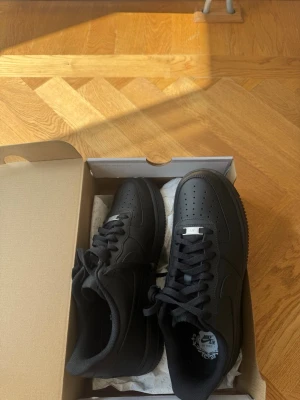 Nike Air Force 1 helsvarta sneakers - Säljer ett par helsvarta Nike Air Force 1 sneakers i klassisk low-modell. Skorna har ovandel i slätt läder, perforerad tåbox och svart sula. Ikonisk Nike-logga på sidan och tungan. Perfekta för dig som vill ha en clean och tidlös look. Helt oanvända!