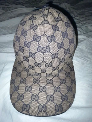 Beige blå röd keps med GG-mönster från Gucci - Säljer en beige blå röd keps från Gucci med det klassiska GG-monogrammönstret över hela kepsen. På sidan finns en ikonisk röd och marinblå rand. Kepsen har böjd skärm och justerbar rem bak. Materialet är canvasliknande tyg. Mycket bra skick Storlek Xs. Limited edition 