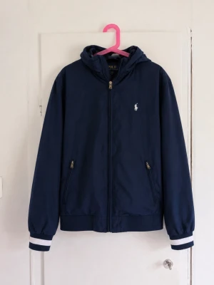 Polo Ralph Lauren jacka marinblå – helt ny - Helt ny Polo Ralph Lauren-jacka i marinblått 💙 Klassisk och stilren modell med huva, vit broderad Polo-logga på bröstet och snygga vit/marinblå detaljer vid ärmsluten. Två fickor med dragkedja framtill och full dragkedja. • Storlek L/G (14–16) • Helt ny / oanvänd • Marinblå • Huva • Broderad Ralph Lauren-logga • Väldigt fint skick – inga anmärkningar Perfekt som vår-, höst- eller lättare sommarjacka. Skriv gärna vid frågor eller om du vill ha mått! 💙