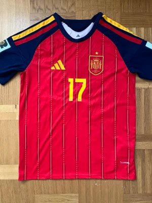 Spaniens matchtröja Adidas röd - Säljer en röd matchtröja från Adidas för Spaniens landslag med gula detaljer, nummer 17 på bröstet och landslagets emblem. Tröjan har mörkblå ärmar med gula ränder och är tillverkad i lätt funktionsmaterial. Perfekt för fotbollsträning eller att bära på läktaren.