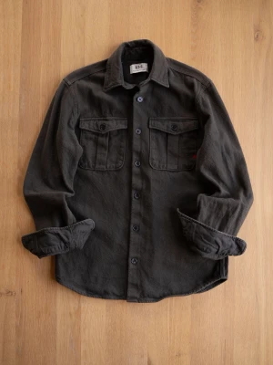 Mörkgrå overshirt med fickor - Stilren mörkgrå overshirt med två bröstfickor med lock och knappar. Skjortan har klassisk krage, knäppning framtill och långa ärmar. Materialet ser ut att vara mjukt och lite tjockare, perfekt för lager-på-lager. Snygg detalj med liten röd etikett vid ena fickan.