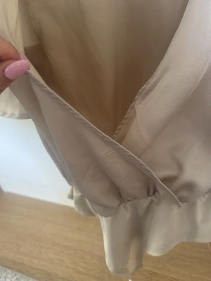 Beige omlottopp från NA-KD - Säljer en beige omlottopp från NA-KD i storlek 38. Toppen har en djup v-ringning och omlottdesign med resår i midjan för en snygg passform. Perfekt att styla med jeans eller kjol för en trendig look.