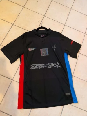 Svart fotbollströja Cactus Jack x Nike - Säljer en svart fotbollströja från Nike i samarbete med Cactus Jack. Tröjan har röda och blå detaljer på sidorna, tryck på bröstet och ärmen samt grafiska prints framtill. Materialet är troligtvis polyester och modellen har korta ärmar och V-ringning.