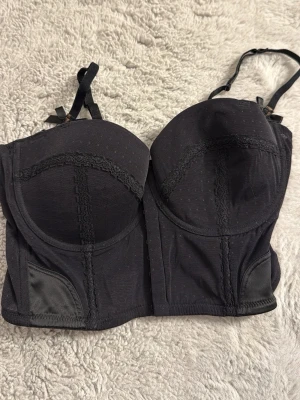 Svart bustier-bh med spetsdetaljer - Snygg svart bustier-bh med vadderade kupor och dekorativa spetsdetaljer. Justerbara axelband och hakknäppning bak. Materialet är en mix av mesh och tyg med små prickar, vilket ger en lyxig känsla. Perfekt för dig som vill ha något extra under kläderna.