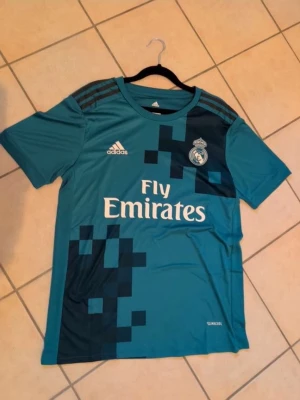 Real Madrid Adidas turkos matchtröja - Snygg turkos matchtröja från Real Madrid med vita loggor och Fly Emirates-tryck på bröstet. Tröjan har mörkare grafiska detaljer, rund hals och klassiska Adidas-ränder på axlarna. Tillverkad i lätt och funktionellt material, perfekt för fotbollsträning.