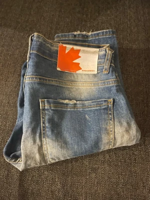 Blå slitna jeans från Dsquared2 - Säljer ett par blå jeans från Dsquared2 med slitningar, hål och färgstänk för en riktigt edgy look. Jeansen har en klassisk femficksmodell och en stor lapp med röd lönnlöv på bakfickan. Perfekta för dig som gillar streetstyle och statement-plagg.