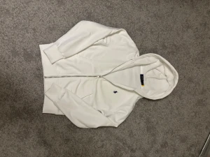 Vit hoodie från Polo Ralph Lauren - Snygg vit hoodie med dragkedja från Polo Ralph Lauren. Klassisk design med huva, ribbade muddar och liten broderad logga på bröstet. Perfekt för en clean och stilren look. Tillverkad i mjukt material för extra komfort. Skulle REKOMMENDERA till nån som har storlek XS