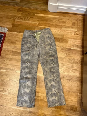 Ormmönstrade raka jeans - Säljer ett par unika raka jeans med ormmönster i grå och beige toner. Byxorna har hög midja och dragkedja framtill. Materialet känns mjukt och har en lätt glansig finish. Perfekta för dig som vill sticka ut med din stil.