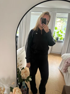 Svart Adidas ziptröja med vita ränder - Klassisk svart ziptröja från Adidas med vita ränder längs ärmarna och Adidas-logga på bröstet. Tröjan har dragkedja framtill, ribbade muddar och två fickor. Perfekt för en sportig och avslappnad stil.
