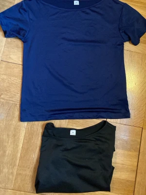 Två basic t-shirts i blått och svart - Säljer två stilrena basic t-shirts i marinblått och svart. Båda har rund halsringning och korta ärmar. Perfekta att matcha till jeans eller shorts för en enkel och clean look.