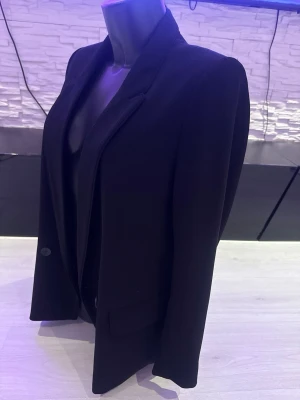 Svart klassisk kavaj med enkelknäppning - Stilren svart kavaj med enkelknäppning och två framfickor. Kavajen har smal krage och lång ärm, vilket ger en clean och tidlös look. Passar perfekt till både jeans och kostymbyxor. Materialet ser ut att vara en mjuk och följsam textil.