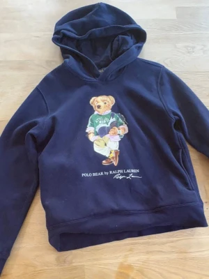 Mörkblå hoodie Polo Bear Ralph Lauren - Mörkblå hoodie med huva från Ralph Lauren, prydd med den ikoniska Polo Bear-figuren på bröstet. Trycket visar en nalle i preppy outfit och texten 'POLO BEAR BY RALPH LAUREN'. Hoodien har klassisk passform och mjukt material, perfekt för en avslappnad stil.