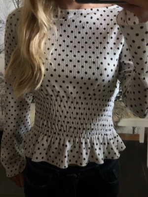 Vit prickig blus med smock - Supersöt vit blus med svarta prickar och smockad. Blusen har långa ärmar och volang. Använd endast fåtal gånger