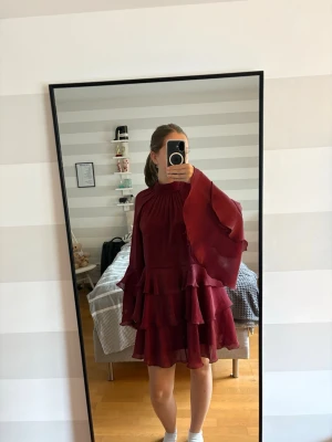 Vinröd volangklänning med vida ärmar - Supersnygg vinröd kortklänning med volanger och vida, fladdriga ärmar. Hög hals och flera lager volang i kjolen ger en riktigt trendig look.Perfekt för dig som vill sticka ut lite extra. Materialet känns lätt och flowy. 