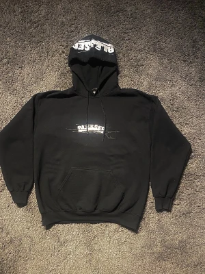 Svart hoodie med Blessed-tryck - Säljer en svart hoodie med stor huva och magficka. På bröstet finns ett vitt tryck med texten 'BLESSED' i molnliknande stil och samma tryck återfinns på huvan. Hoodien har ribbade muddar och är gjord i mjukt material, perfekt för en avslappnad streetwear-look.