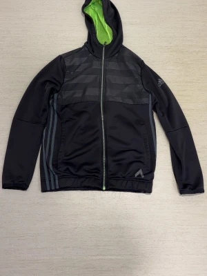 Svart hoodie från Adidas med dragkedja - Svart hoodie från Adidas med hel dragkedja och huva med grön insida. Snygga gråa ränder på sidorna och diskret mönster upptill. Adidas-logga på ärmen. Perfekt för en sportig och avslappnad stil.