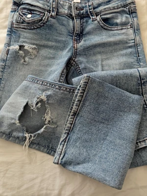 Ljusa slitna jeans från Gina Tricot - Säljer ett par ljusblå jeans från Gina Tricot i storlek 164. Jeansen har trendiga slitningar och hål på ena benet samt klassisk femficksmodell. Materialet är denim med snygg tvätt och kontrastsömmar. Perfekta för en avslappnad och cool stil.