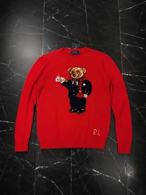  Polo Ralph Lauren - Röd stickad tröja från Polo Ralph Lauren med ikonisk björn i kostym och halsduk på framsidan. Tröjan har rund halsringning, ribbade muddar och RL-broderi nertill. Perfekt för dig som vill ha en unik och lekfull look.
