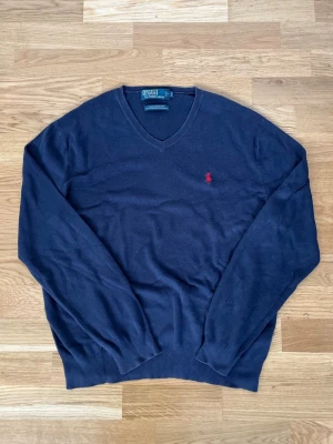 Marinblå stickad tröja Polo Ralph Lauren - Marinblå stickad tröja från Polo Ralph Lauren med klassisk röd logga broderad på bröstet. Tröjan har v-ringning, långa ärmar och ribbade muddar vid ärmslut och nederkant. Perfekt för lager på lager och stilren look. Enda egentliga defekten är vid storlekslappen som sitter lite löst, men ingen som påverkar.