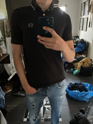 Svart pikétröja ifrån Armani exchange - Snygg svart pikétröja med vit kant på kragen och liten vit logga på bröstet. Tröjan har korta ärmar och knäppning med tre knappar framtill. Perfekt till jeans eller chinos för en clean och stilren look.