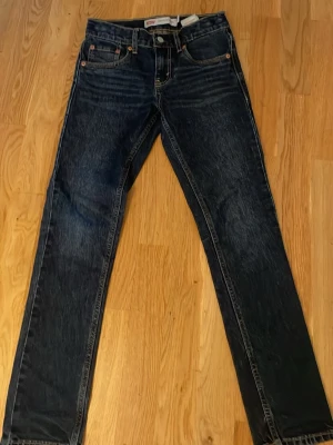 Mörkblå  jeans från Levi's - Säljer ett par klassiska mörkblå jeans från Levi's med raka ben och fem fickor. Jeansen har snygga slitningar och kontrastsömmar samt den ikoniska läderlappen bak i midjan. Perfekta för en avslappnad och tidlös stil.